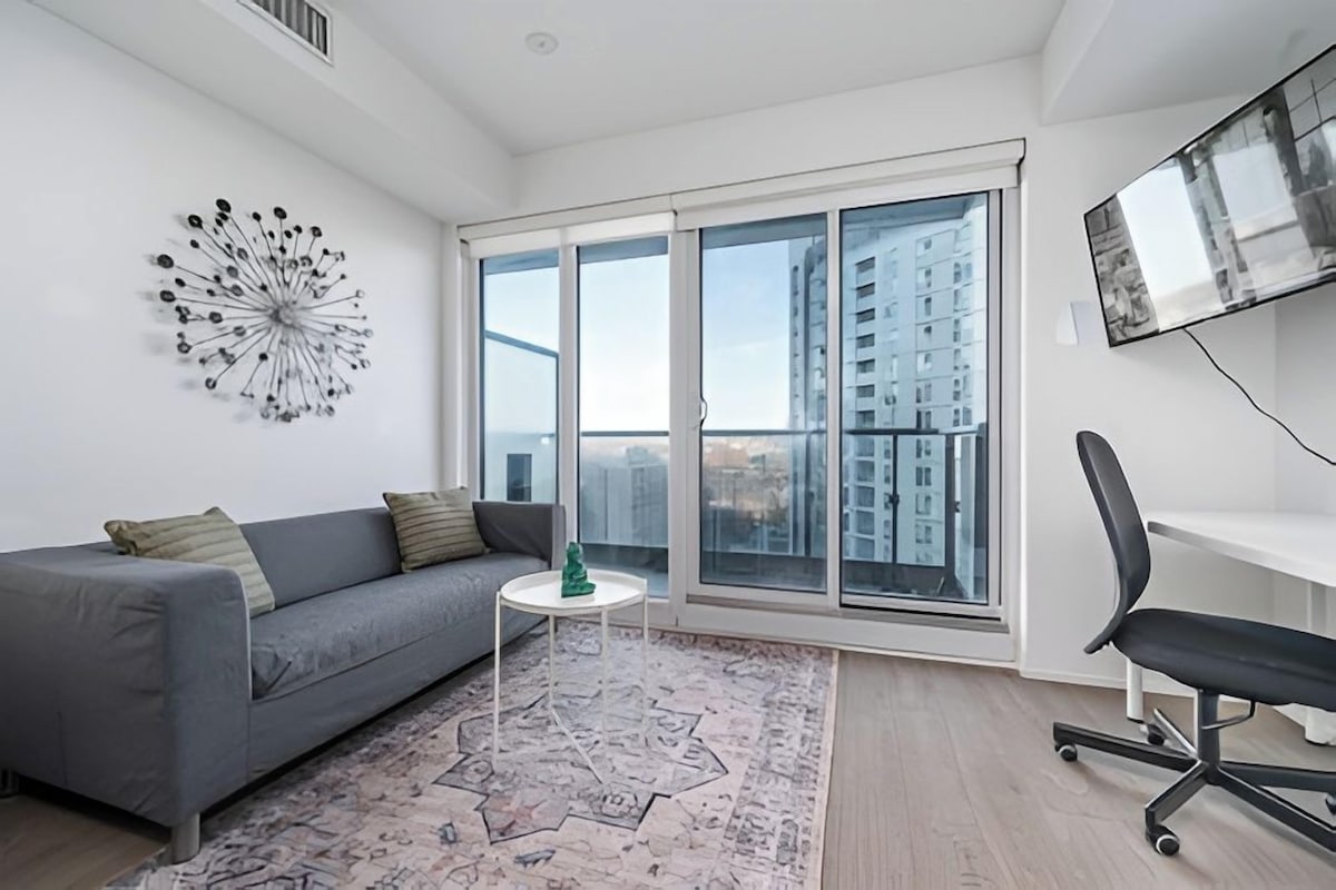 A modern living room with a grey sofa, a small white table, a decorative wall piece, a large window with a view of high-rise buildings, and a mounted TV.