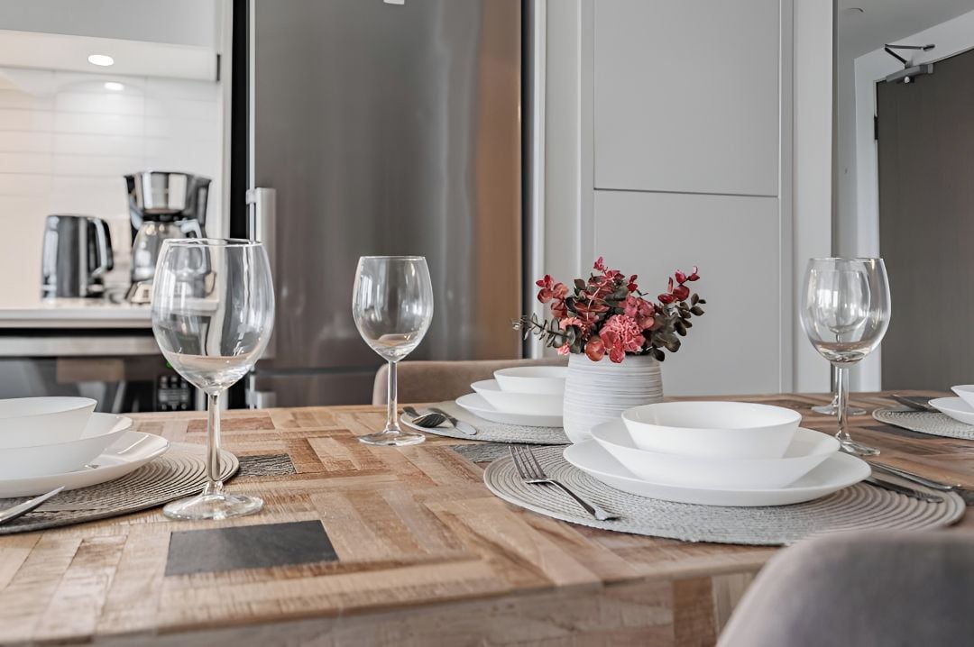 Une table à manger dressée avec de l’assiette blanche, des verres et des couverts, et un vase rempli de fleurs placé au centre.
