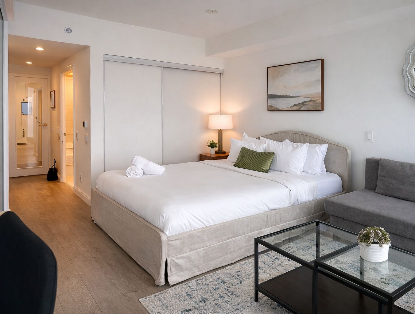 A hotel room with a bed, a sofa, a glass coffee table, a lamp, and a painting on the wall.