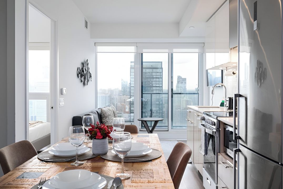 A modern kitchen with stainless steel appliances, a dining area with a wooden table, and large windows offering a city view.