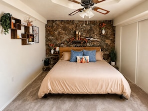A bedroom with a stone wall, a bed with a beige bedspread, a ceiling fan, and a nightstand with a lamp and a vase.