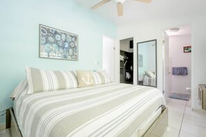 Une chambre à coucher comprenant un lit, un ventilateur de plafond, une porte donnant sur une salle de bain et une œuvre d’art accrochée au mur.