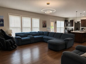 Een moderne woonkamer met een hoekbank, een gaming stoel en een keuken met houten kastjes en een aanrecht.