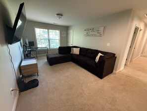 Ein Wohnzimmer mit einer Eckcouch, einem flachbildfernseher an der Wand, einem Schreibtisch mit einem Computer und einem Fenster mit Jalousien.
