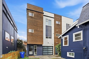A modern multi-story residential building with a combination of wooden and metal siding, a garage, and a small front yard with a recycling bin. - Rooftop Retreat w/ Stunning Views Ballard Gem (Seattle)