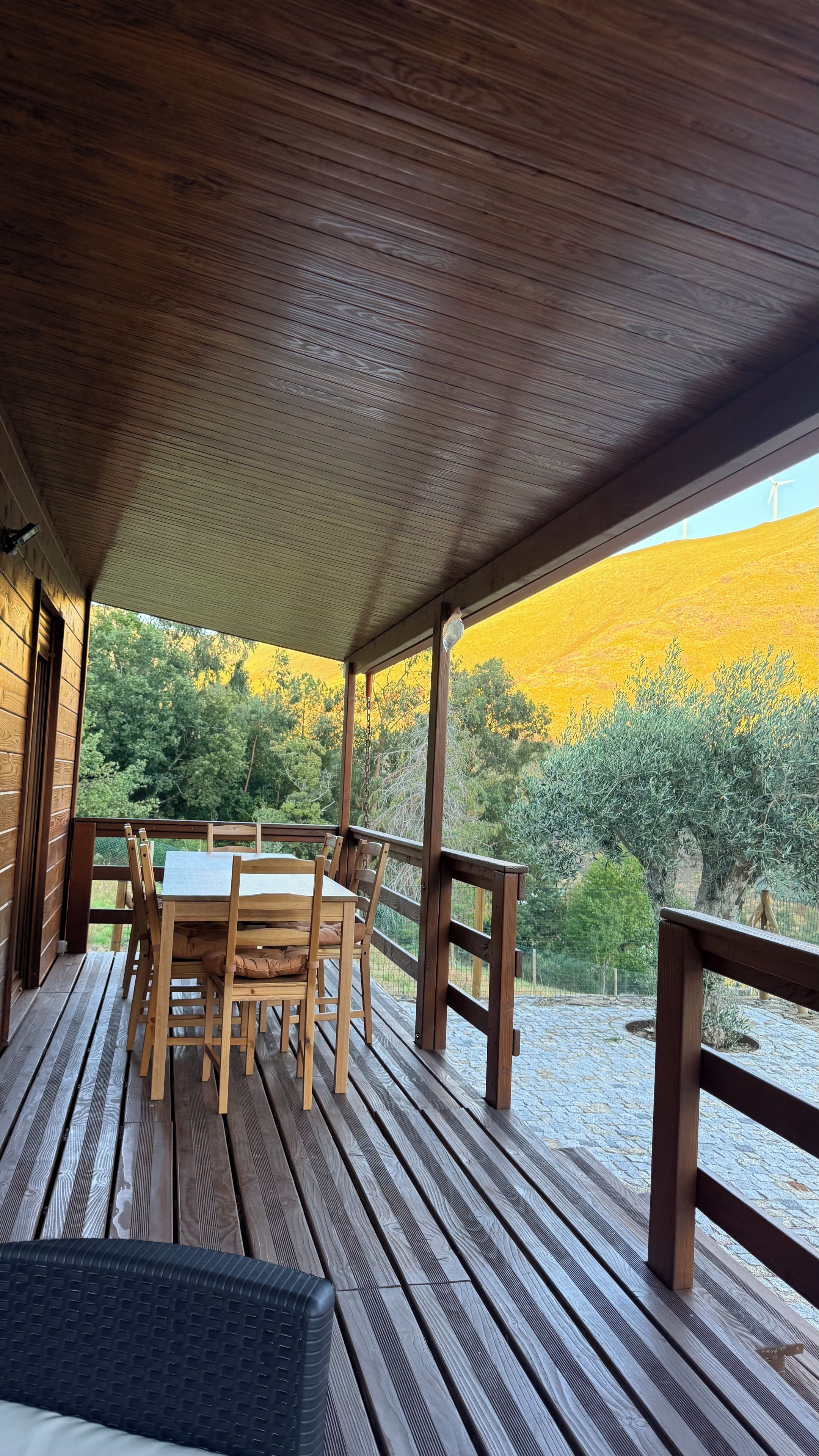 Una terraza de madera con una mesa y sillas, con vista a un paisaje natural con árboles y una colina.