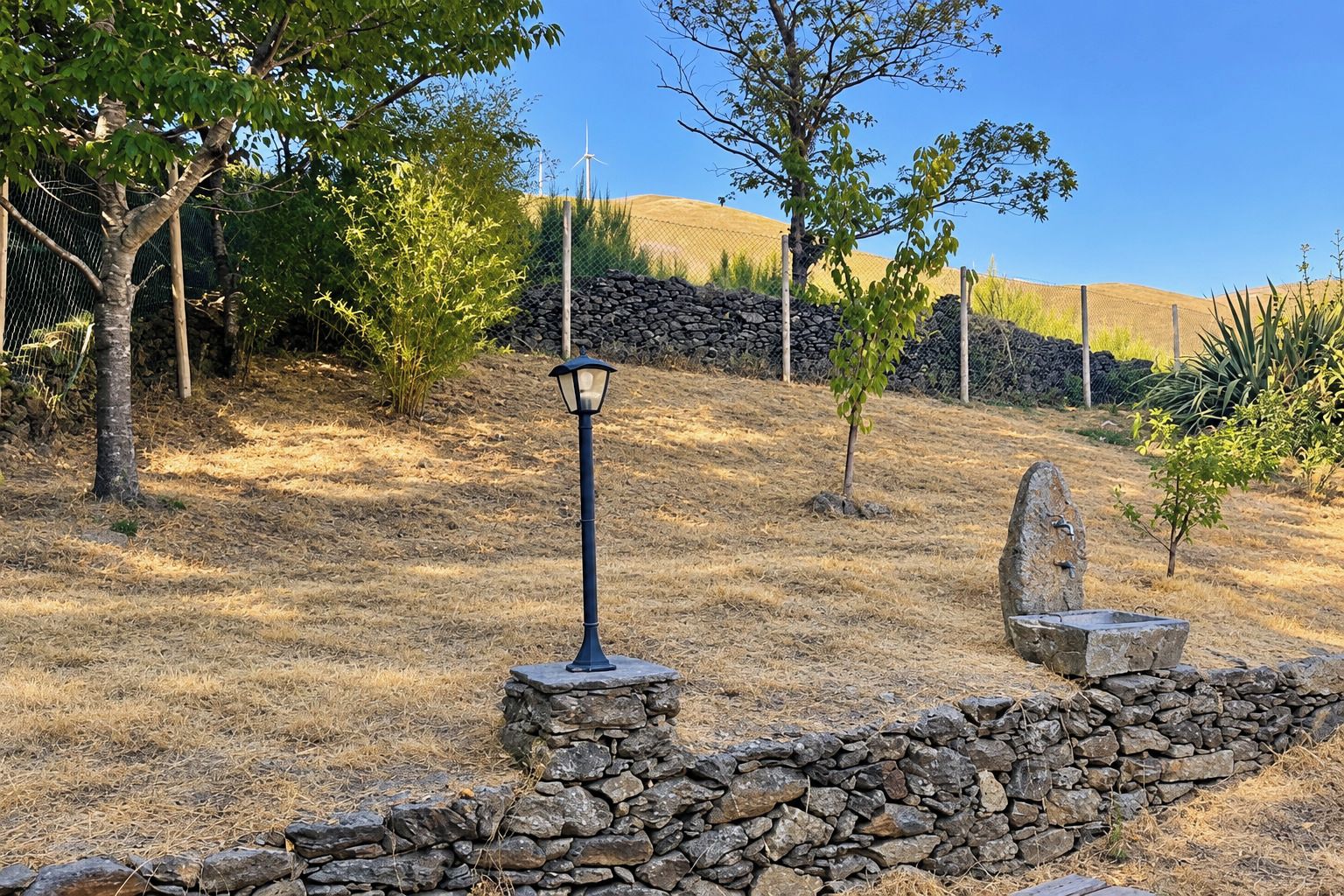 Un muro de piedra con un poste de farol, un pequeño árbol y una estructura de piedra que se asemeja a un pozo o una fuente en un área seca y cubierta de hierba, con árboles y una valla al fondo.
