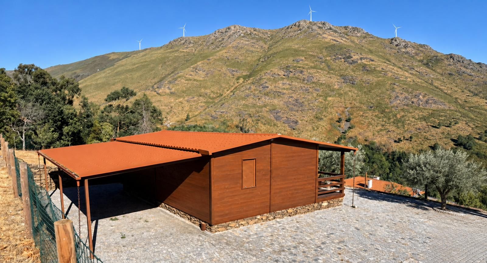 A wooden building with a sloped roof, surrounded by a stone-paved area and a green fence.