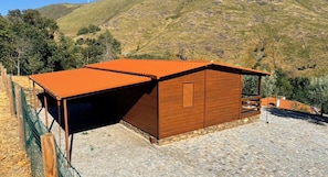 A wooden building with a sloped roof, situated on a stone-paved area with a view of a hilly landscape.