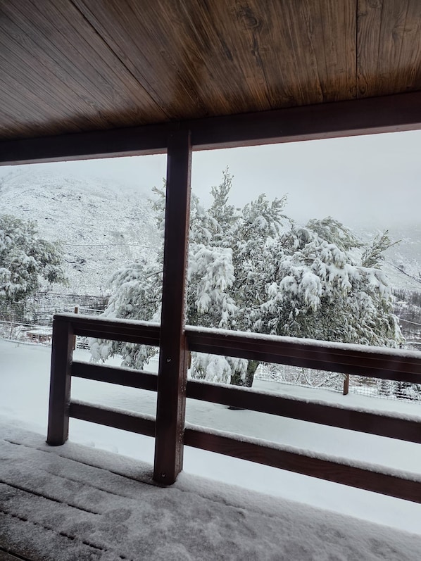 A wooden bench with a metal railing, overlooking a snowy landscape with trees and a hill.