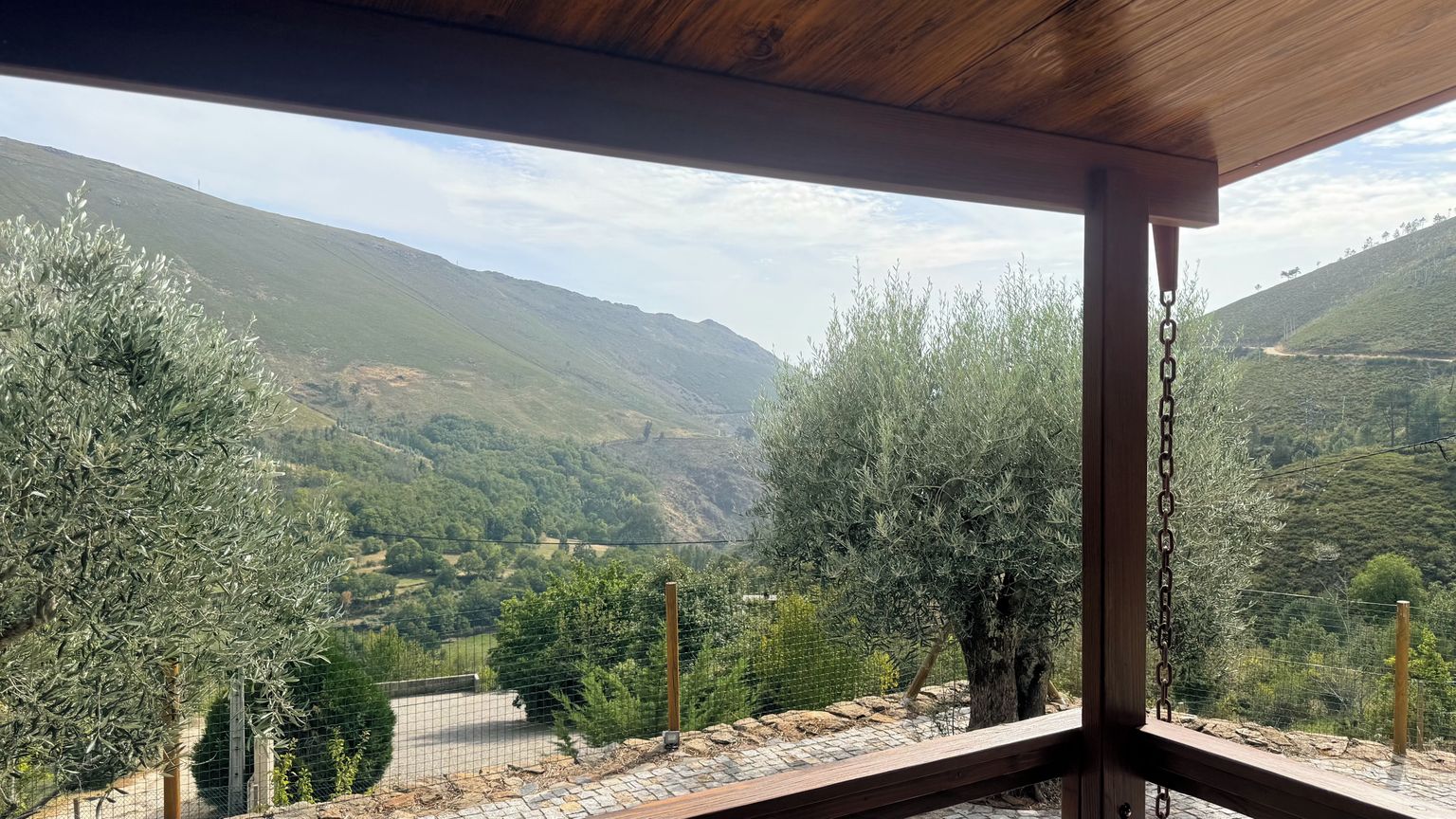 Vue depuis un espace extérieur abrité, doté d’un plafond en bois, donnant sur une vallée plantée d’oliviers et sur une chaîne de montagnes.