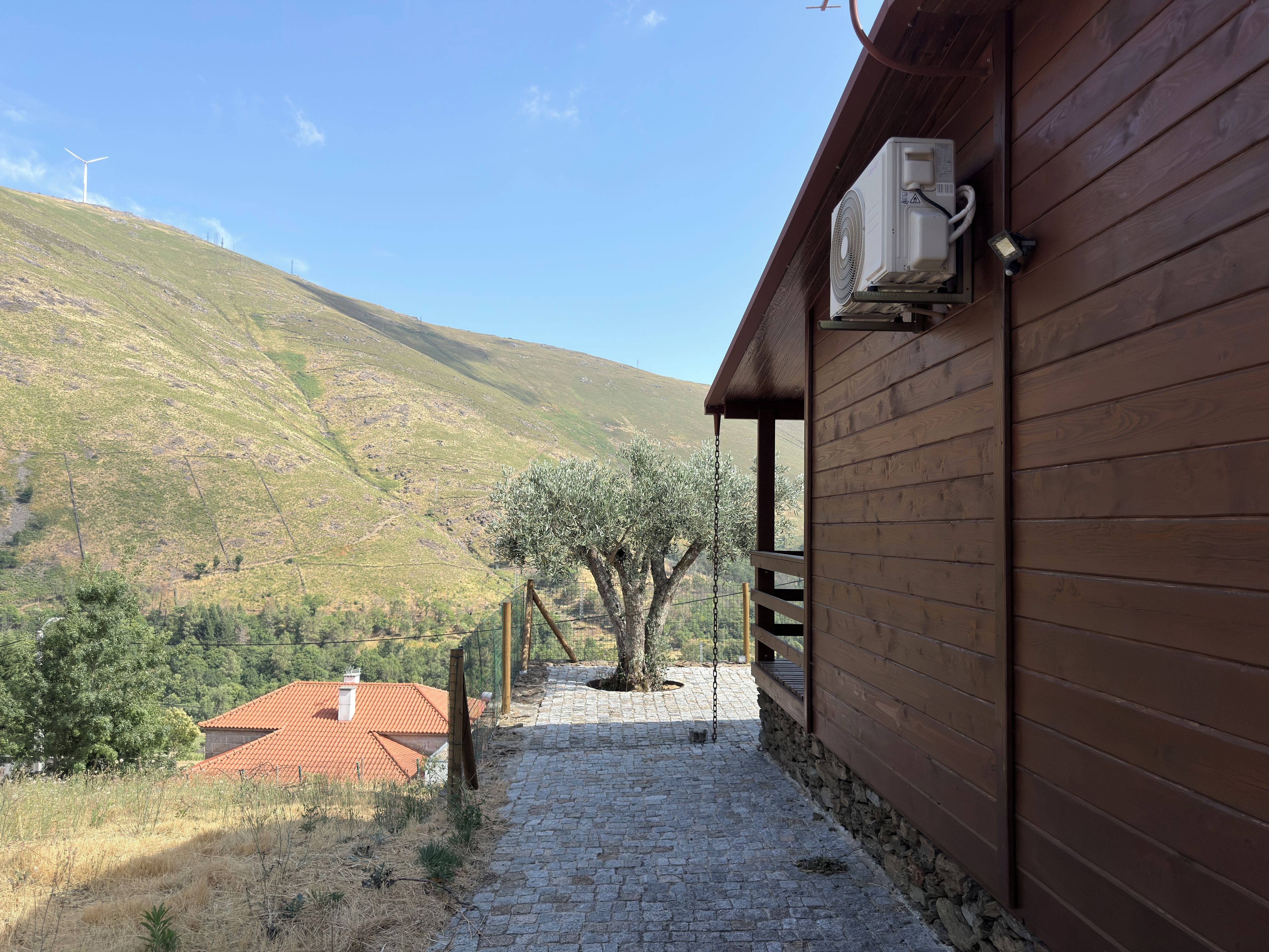 Un edificio con un sendero de piedra que lleva a una puerta de madera, vista de una colina con aerogeneradores y un árbol en primer plano.