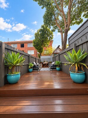 A wooden deck with a seating area, potted plants, and a view of buildings in the background. - Entire 3BR Beachside House in the Heart of St Kilda (St Kilda)