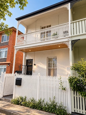 Un immeuble résidentiel de deux étages, doté d’une clôture en bois peinte en blanc, d’un balcon avec des balustrades blanches décoratives et d’une façade en briques.