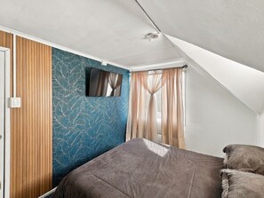A bedroom with a sloped ceiling, a bed with a brown cover, a wall-mounted TV, and a window with sheer curtains.