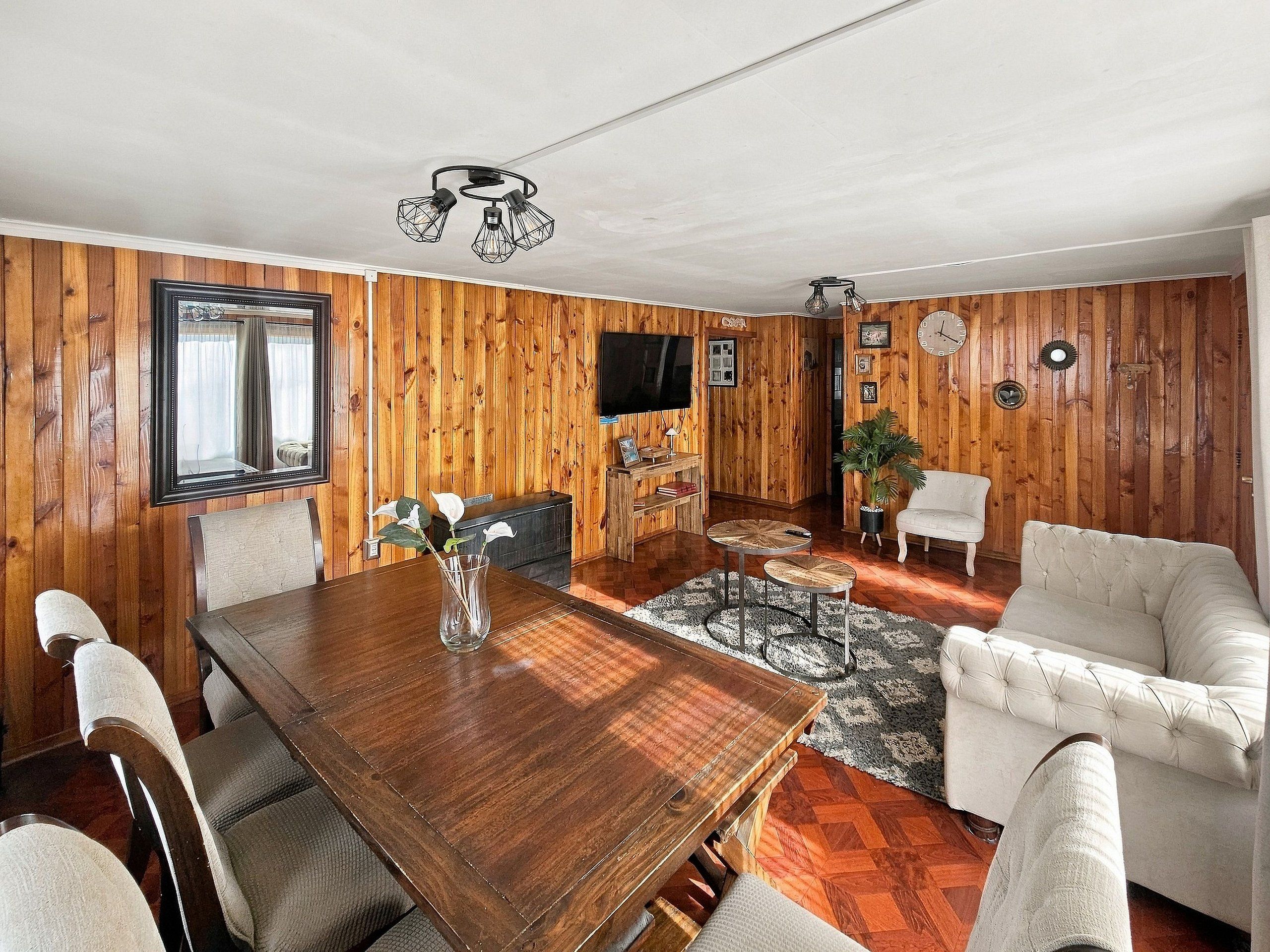 Quarto com parede revestida de madeira, mesa de jantar com cadeiras, sofá, televisão e luminária no teto.