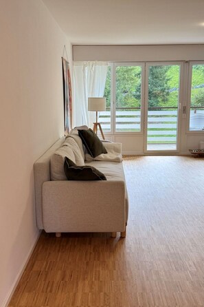 A modern living room with a sofa, a floor lamp, and a view of greenery through large windows. - Waldhaus A1/4 (CharmingStay), (Flumserberg Tannenboden), by Interhome (Flumserberg Tannenheim)