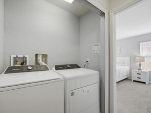 Ein Waschraum mit zwei Frontlader-Waschmaschinen und -Trocknern, ein kleiner, weißer Nachttisch mit Lampe sowie ein Bett im angrenzenden Zimmer.