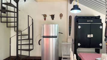 A compact kitchen with a stainless steel refrigerator, a black cabinet with decorative handles, a wooden staircase, and a radiator.