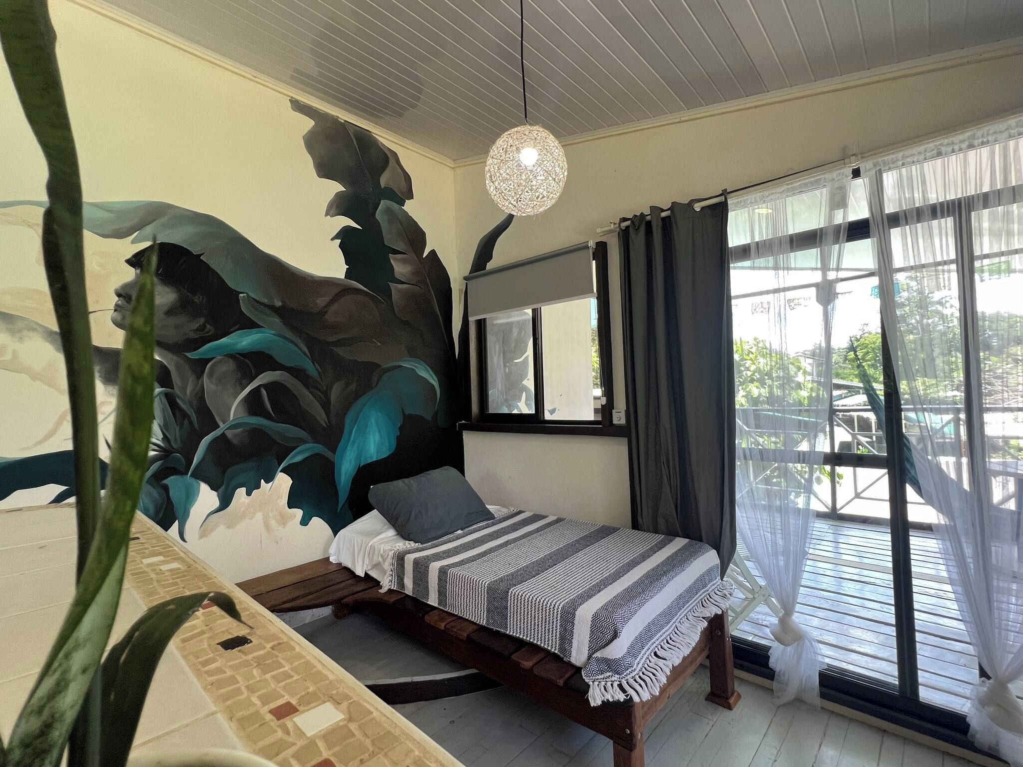 A bedroom with a bed, a large window with curtains, a hanging light fixture, and a wall mural.