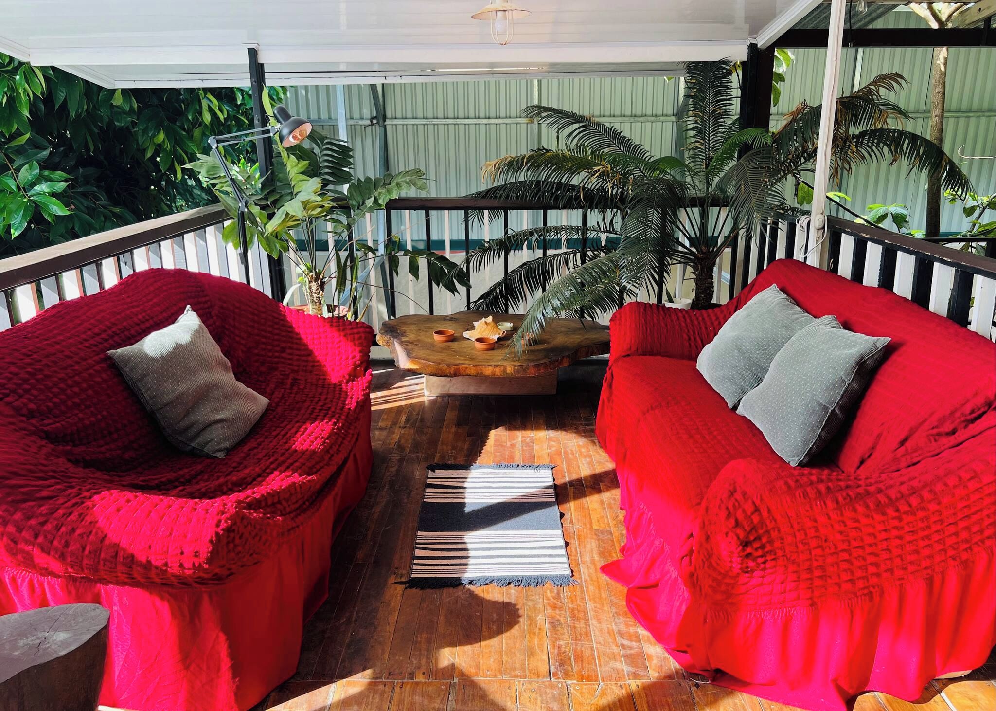 A covered outdoor seating area with two red cushioned chairs and a wooden table, surrounded by potted plants.