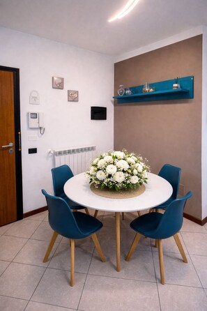 Uma área de jantar com uma mesa redonda, cadeiras azuis, um arranjo floral, uma prateleira na parede e uma porta com tranca.