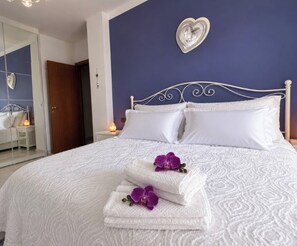 A neatly made bed with white linens and purple flowers, a heart-shaped wall decoration, and a mirror reflecting part of the room. - Strategically located near downtown Milan, Bocconi, San Siro, Assago, and Humanitas. (Rozzano)