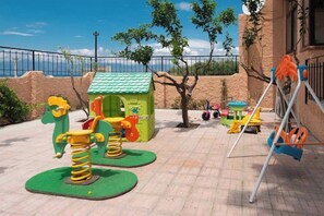 Een speeltuin met diverse speeltoestellen, waaronder schommels en een klimconstructie, met op de achtergrond een heldere lucht en een gebouw.