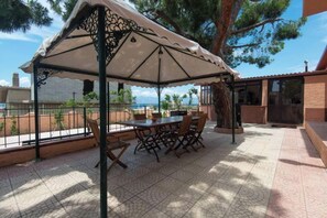 Een terras buiten met een tafel en stoelen onder een overkapping, omringd door een hek en bomen.