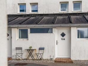 Een gebouw met twee verdiepingen, met een witte gevel, nummer 104, met een klein voortuintje met twee stoelen en een tafel, en een deur met een ruitvormig raam.