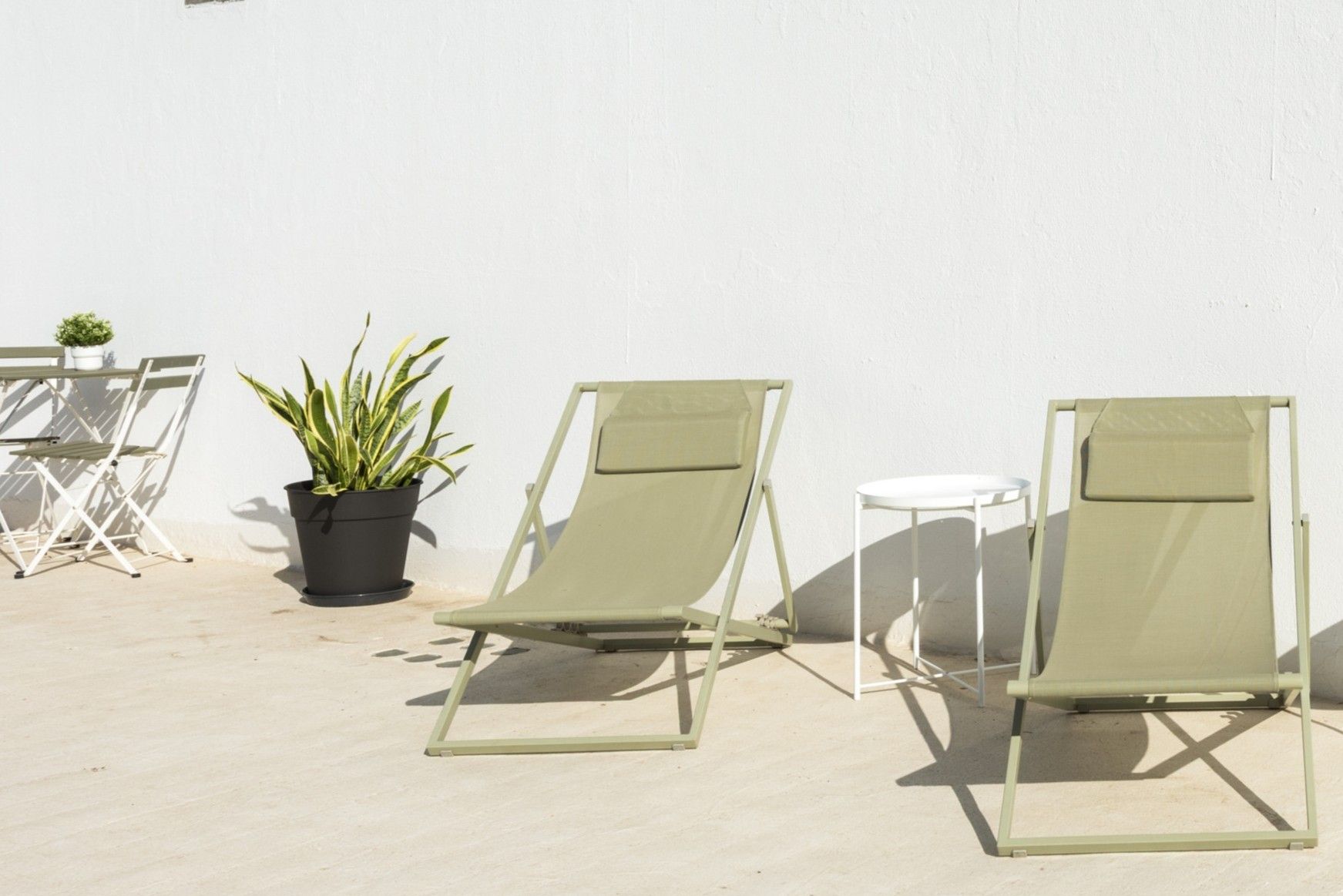 Trois chaises de jardin vertes et une petite table sont placées contre un mur blanc, avec une plante en pot à proximité.