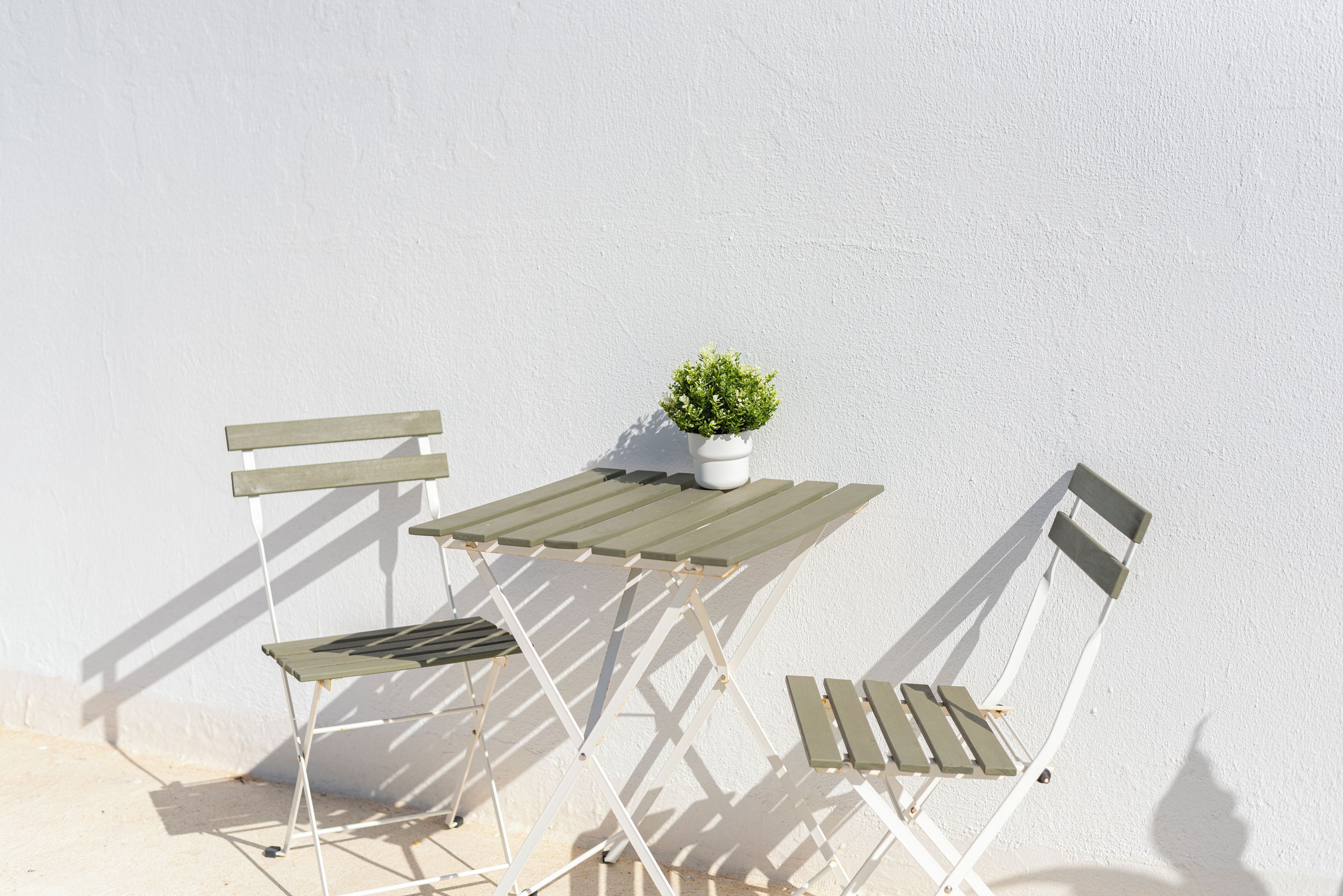 A white outdoor table and two white chairs with wooden slats, a small potted plant on the table, and a white wall in the background.