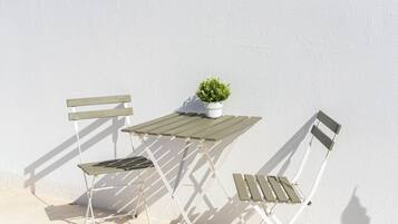 A white outdoor table and two white chairs with wooden slats, a small potted plant on the table, and a white wall in the background.