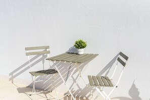 A white outdoor table and two white chairs with wooden slats, a small potted plant on the table, and a white wall in the background.