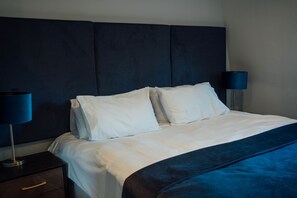 Ein ordentlich bezogenes Bett mit weißen Leinen und einem dunkelblauen Kopfteil. An beiden Seiten des Bettes stehen Nachttischlampen mit blauen Lampenschirmen.
