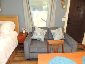 Ein Schlafzimmer mit einem Bett, einem grauen Sofa mit gemusterten Kissen, einem Holfnachtkästchen und einem Holzstuhl.