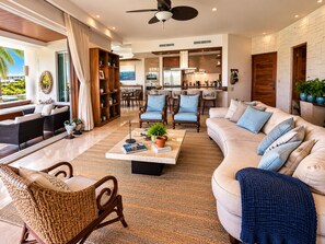 A spacious living area with a sofa, chairs, a coffee table with books and a plant, a dining area with chairs and a kitchen in the background. - Luxury Peninsula Condo with Canal and Ocean Views / Newly Renovated / 3 Bedrooms (Puerto Aventuras)