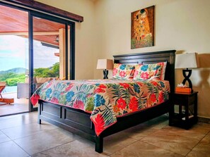 A bedroom with a bed, a nightstand, a painting, and a view through a glass door. - Casa de Shanti: Private Ocean-View 4BR Retreat with Infinity Pool & Hot Tub (Coco)