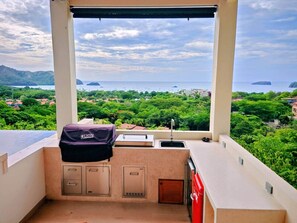 A balcony with a barbecue grill, sink, and cabinets, overlooking a lush landscape and the sea. - Casa de Shanti: Private Ocean-View 4BR Retreat with Infinity Pool & Hot Tub (Coco)
