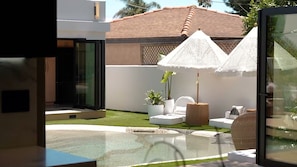 Ein moderner Außenbereich mit einem Glastisch, weißen Lounge-Stühlen und einem kleinen Garten mit weißem Zaun und Topfpflanzen.