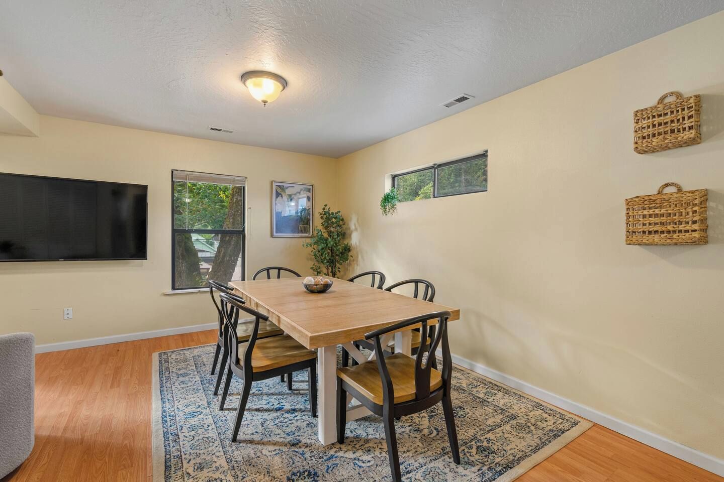 A dining area with a wooden table, chairs, a rug, and a television mounted on the wall.