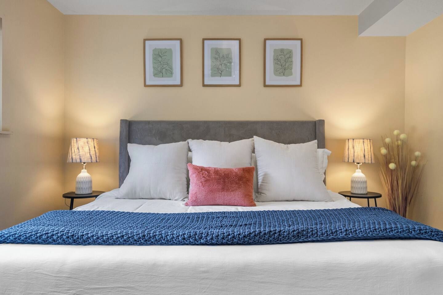A hotel room with a bed, two bedside lamps, a headboard, and three framed botanical prints on the wall.