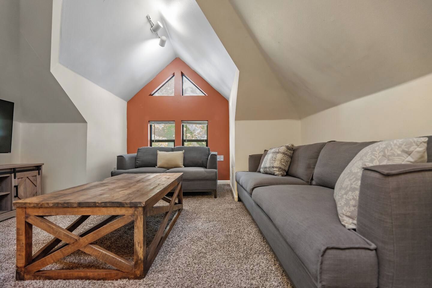 A loft-style living room with a sloped ceiling, a wooden coffee table, two grey sofas, and a television on the left.