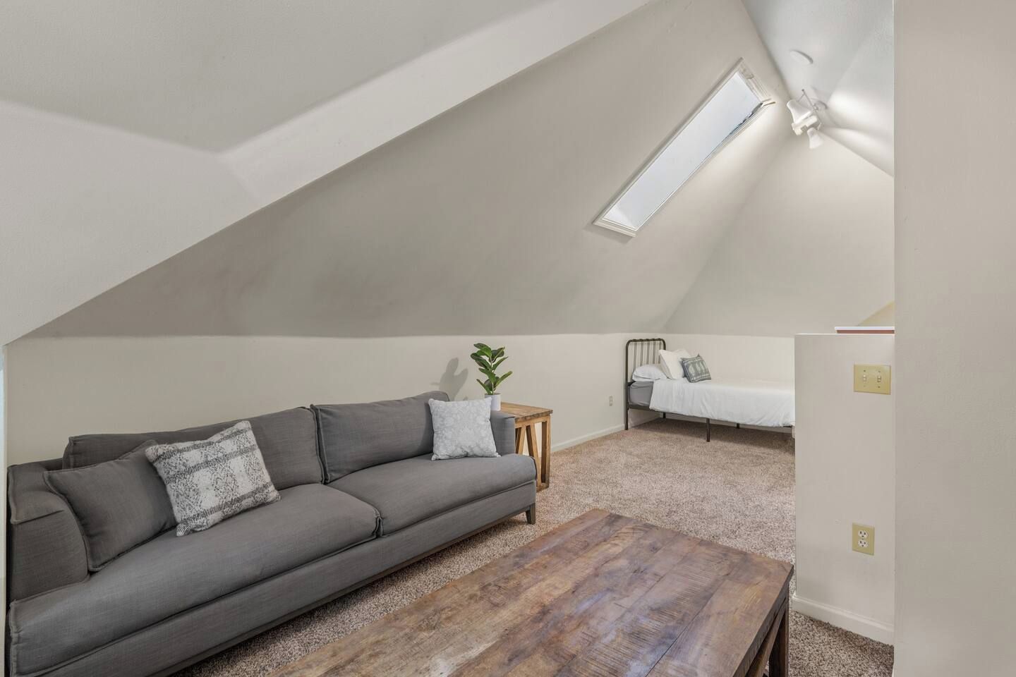 A loft bedroom with a sloped ceiling, a gray sofa, a wooden coffee table, a small bedside table, and a bed.