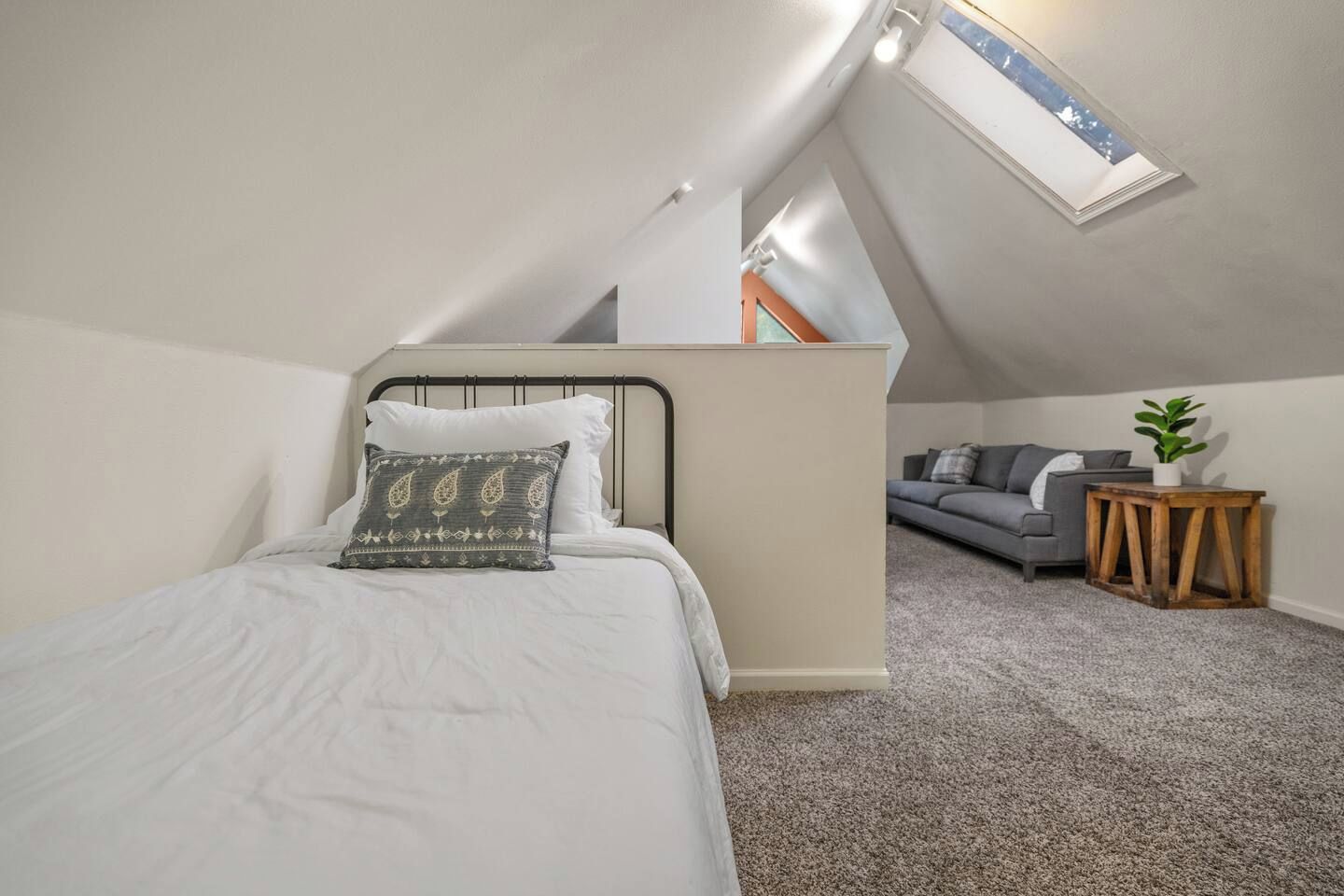 A bedroom with a sloped ceiling, a bed with a patterned pillow, a grey sofa, and a small wooden table with a plant.