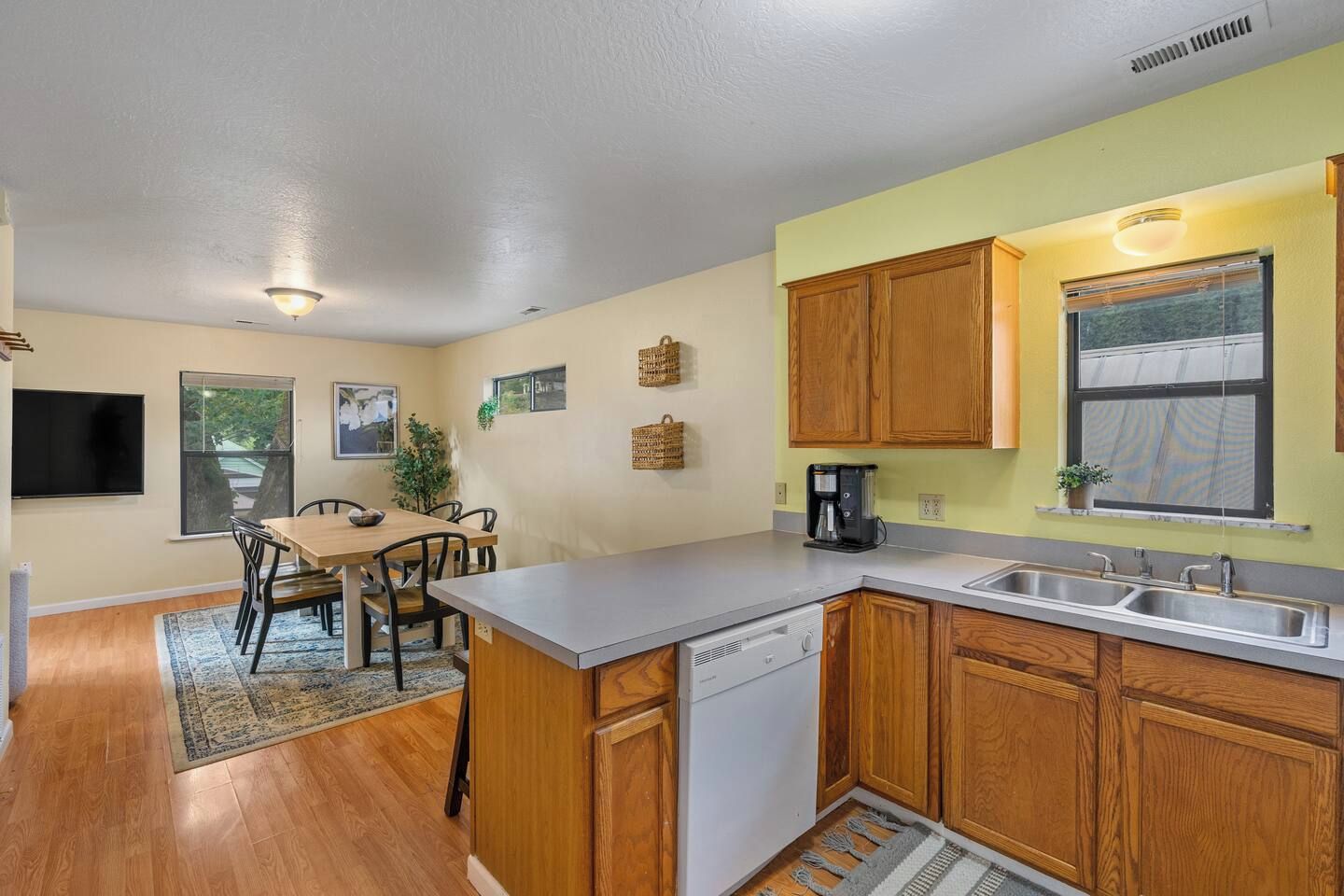 A compact kitchen with wooden cabinets, a built-in dishwasher, a coffee maker, and a window with blinds.