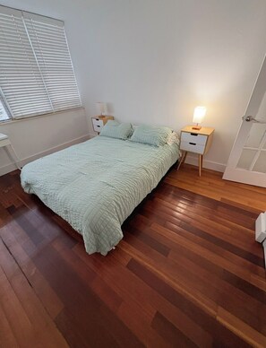 Ein Schlafzimmer mit einem Bett, zwei Nachttischen, einer Lampe und einem Fenster mit Jalousien.