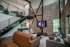 A spacious living area with a high ceiling, a brick wall, a staircase, and a television mounted on the wall. - "Rest & Redeem" New Luxury Cabin in Broken Bow (Broken Bow)