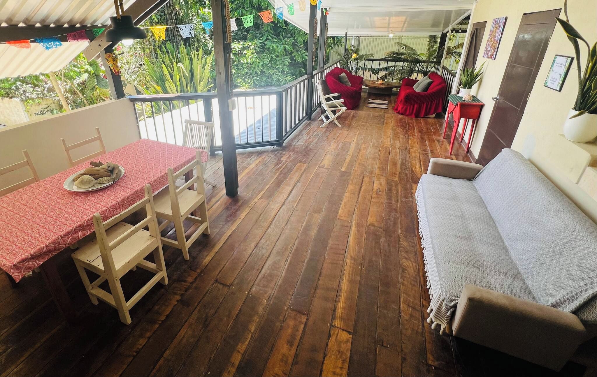 Un espacio al aire libre techado con piso de madera, una mesa de comedor con sillas, un sofá y una barandilla.