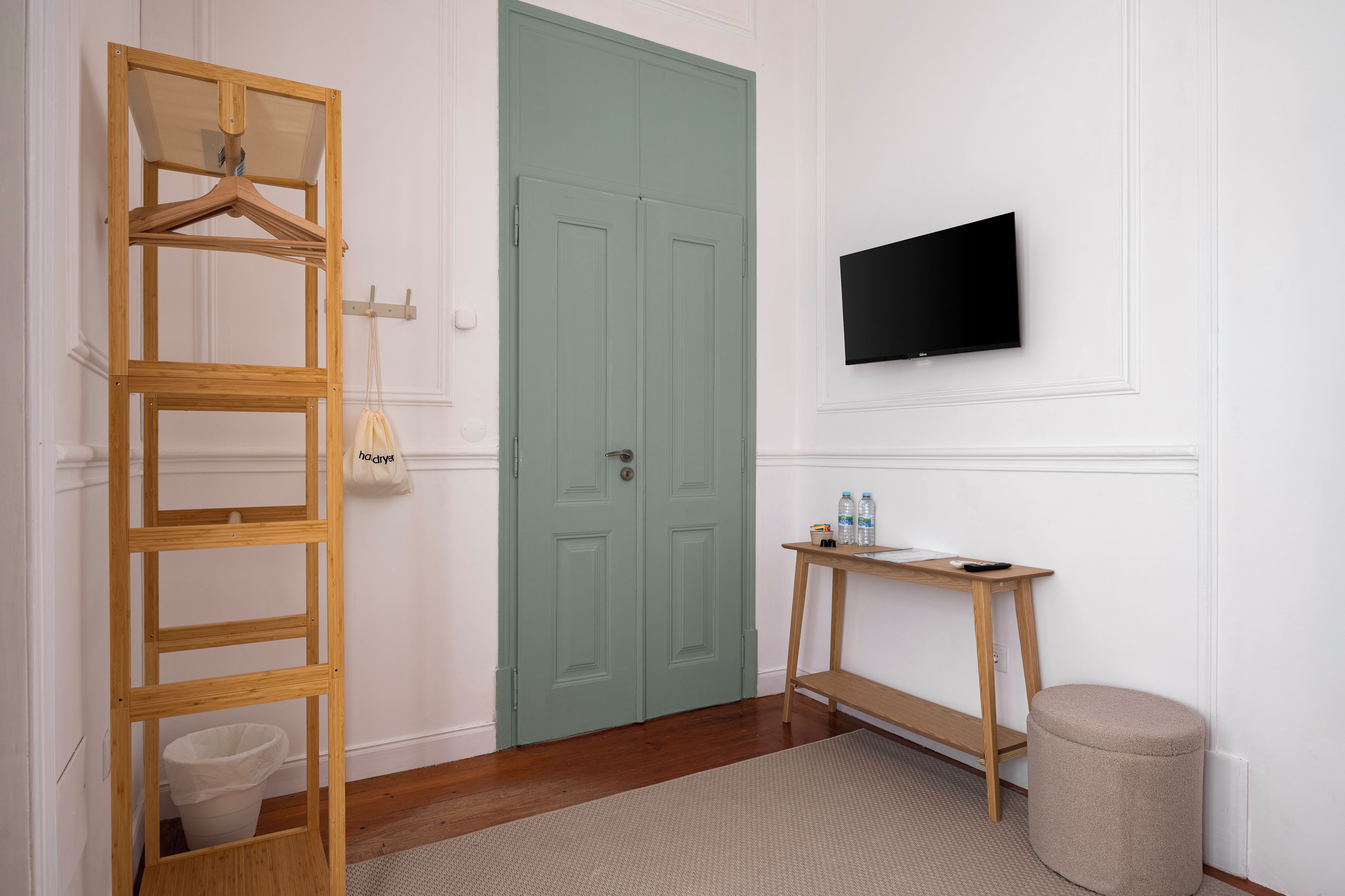 Une pièce avec une échelle en bois, une porte verte, un téléviseur fixé au mur, une petite table en bois avec des bouteilles et un pouf rond.